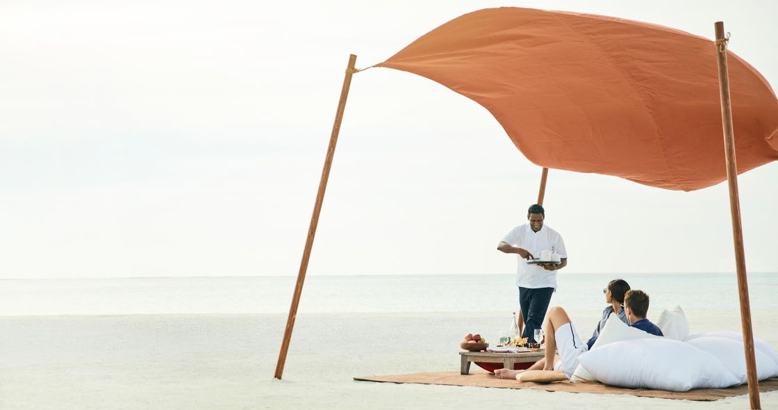 A Man Standing Next To A Couple Of Women Sitting Under An Umbrella