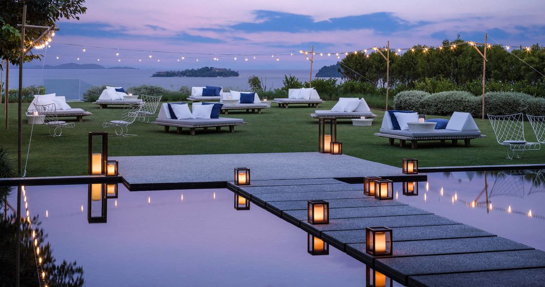 A Pool With A Deck And Chairs And A Body Of Water With Trees And A Sunset