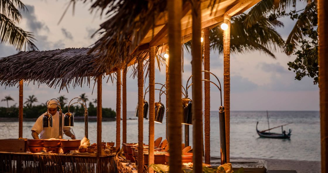 A Person Sitting At A Table Under A Palm Tree On A Beach